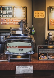 A retro-style Faema E61 espresso machine is prominently displayed on a counter, with informational plaques beside it. The background features a brown wall with various informational panels and a small Brazilian flag. A sign with the words 'COFFEE CUPPER' is visible along with other coffee-related paraphernalia.
