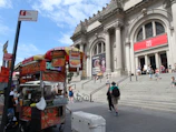 A food cart selling items like hot dogs and drinks is situated on the sidewalk near the entrance of a grand, classical building with columns and steps, identified as 'The Met' by the banner. Several people are visible on the steps and around the cart, with a clear blue sky above.