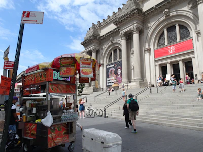 A food cart selling items like hot dogs and drinks is situated on the sidewalk near the entrance of a grand, classical building with columns and steps, identified as 'The Met' by the banner. Several people are visible on the steps and around the cart, with a clear blue sky above.
