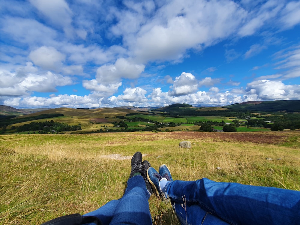 a person's legs and feet on a grassy hill with trees and blue sky