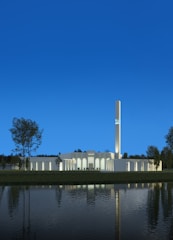 A modern architectural building with a tall, illuminated tower, surrounded by trees and reflected in a calm water body under a clear blue sky.
