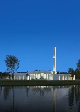 A modern architectural building with a tall, illuminated tower, surrounded by trees and reflected in a calm water body under a clear blue sky.