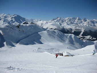 Skiers glide down a snowy slope with expansive views of snow-covered mountains in the background. The clear blue sky enhances the serene and cold atmosphere. Small figures can be seen in the distance enjoying the vast ski area.
