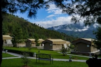 A series of tan canvas tents are set up in a lush, green campsite surrounded by pine trees and rolling hills. The tents have a rustic appearance and are arranged neatly with stone pathways leading to them. A decorative bench is placed in the foreground. The sky is partly cloudy, with white clouds hovering over the distant mountains.