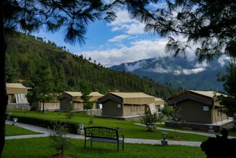 A series of tan canvas tents are set up in a lush, green campsite surrounded by pine trees and rolling hills. The tents have a rustic appearance and are arranged neatly with stone pathways leading to them. A decorative bench is placed in the foreground. The sky is partly cloudy, with white clouds hovering over the distant mountains.