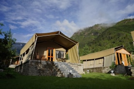 A group of luxury tents with canvas roofs and wooden frames, set on stone platforms with steps leading up to the entrance. The setting is surrounded by lush green grass and tall trees, with a backdrop of mist-covered mountains and a partly cloudy sky.
