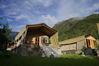 A group of luxury tents with canvas roofs and wooden frames, set on stone platforms with steps leading up to the entrance. The setting is surrounded by lush green grass and tall trees, with a backdrop of mist-covered mountains and a partly cloudy sky.