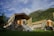 A group of luxury tents with canvas roofs and wooden frames, set on stone platforms with steps leading up to the entrance. The setting is surrounded by lush green grass and tall trees, with a backdrop of mist-covered mountains and a partly cloudy sky.
