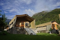 A group of luxury tents with canvas roofs and wooden frames, set on stone platforms with steps leading up to the entrance. The setting is surrounded by lush green grass and tall trees, with a backdrop of mist-covered mountains and a partly cloudy sky.
