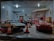 A miniature garage scene with model cars and figures. Several model cars are parked inside a scaled-down garage, while small figures, likely representing people, are positioned around the cars. Orange traffic cones are placed on the ground, and there are various details like posters and signs on the walls, adding realism to the diorama.