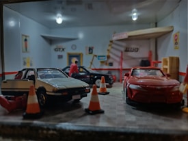A miniature garage scene with model cars and figures. Several model cars are parked inside a scaled-down garage, while small figures, likely representing people, are positioned around the cars. Orange traffic cones are placed on the ground, and there are various details like posters and signs on the walls, adding realism to the diorama.