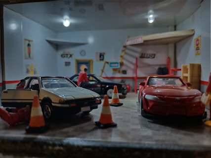 A miniature garage scene with model cars and figures. Several model cars are parked inside a scaled-down garage, while small figures, likely representing people, are positioned around the cars. Orange traffic cones are placed on the ground, and there are various details like posters and signs on the walls, adding realism to the diorama.