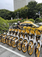 A row of yellow and white bicycles is parked in an organized manner alongside a city street. In the background, a taxi with a matching yellow color is present. A person wearing a helmet is on a motorbike next to the taxi. Lush green trees and a high-rise building are visible, suggesting an urban environment.