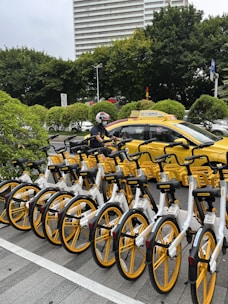 A row of yellow and white bicycles is parked in an organized manner alongside a city street. In the background, a taxi with a matching yellow color is present. A person wearing a helmet is on a motorbike next to the taxi. Lush green trees and a high-rise building are visible, suggesting an urban environment.
