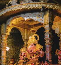 A vibrant and intricate statue of Ganesha is situated inside a richly decorated temple-like structure. The statue is adorned with a gold crown and lavish jewelry, surrounded by ornate columns and floral patterns. Warm lighting enhances the detailed carvings and embellishments.