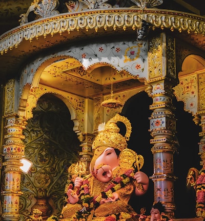 A vibrant and intricate statue of Ganesha is situated inside a richly decorated temple-like structure. The statue is adorned with a gold crown and lavish jewelry, surrounded by ornate columns and floral patterns. Warm lighting enhances the detailed carvings and embellishments.