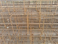 A dense and intricate arrangement of stacked construction rebar shows a pattern of intersecting lines and grids. The rusted metal bars are layered horizontally and vertically, creating a maze-like structure with a uniform, industrial look.