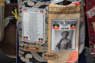 A collage of various cloth patches featuring messages about Australian history and social justice. One patch includes an Aboriginal flag and the phrase 'Fighting Terrorism Since 1770' with an image of an Aboriginal person. Another patch lists various historical massacres in Australia under 'Lest We Forget'. There is also a piece with the text 'Australia was not discovered, it was taken.'