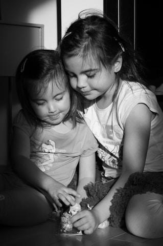 Two young children are sitting closely together on the floor, engaged in play. The scene is illuminated by soft natural light, casting shadows and gently highlighting their expressions of concentration and contentment. They are holding a small toy and seem to be enjoying a quiet moment together.