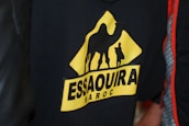 A black garment featuring a triangular yellow logo with the silhouette of a camel and a person. The text 'Essaouira Maroc' is displayed prominently in bold letters below the figures.