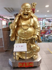 A large golden statue of a cheerful, laughing Buddha is prominently displayed in a shopping mall. The Buddha is adorned with a necklace of large beads and holds a bag in one hand, symbolizing prosperity and contentment. There is a plaque in front of the statue providing information. The surroundings include elements of a shopping environment, with visible stores and advertisements.