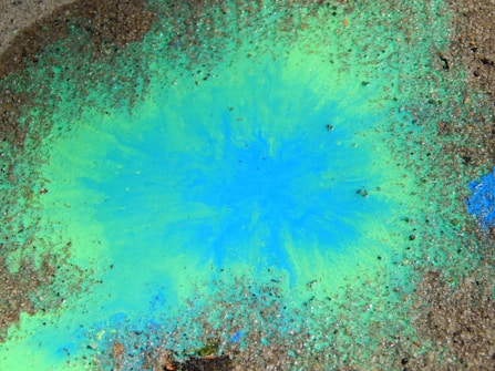 A vibrant splash of blue and green pigments spreads outwards on a rough textured surface, creating a dynamic and organic pattern. The colors blend seamlessly at the edges, while surrounding speckles of what appears to be gravel or sand add an earthy contrast.