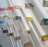Books are displayed in an artful arrangement on a white wall, each book supported by small pegs, and casting shadows that enhance the visual effect. The books have various colored covers, adding vibrancy to the minimalist setting.