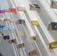 Books are displayed in an artful arrangement on a white wall, each book supported by small pegs, and casting shadows that enhance the visual effect. The books have various colored covers, adding vibrancy to the minimalist setting.