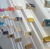 Books are displayed in an artful arrangement on a white wall, each book supported by small pegs, and casting shadows that enhance the visual effect. The books have various colored covers, adding vibrancy to the minimalist setting.
