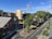 An urban landscape with residential buildings and a road. The area features several apartment complexes with multiple floors, surrounded by lush green trees. The sky is clear with a few clouds, indicating sunny weather. Streets are visible with marked lanes and some traffic cones, suggesting roadworks or restrictions. The scene is calm with no visible people or vehicles.