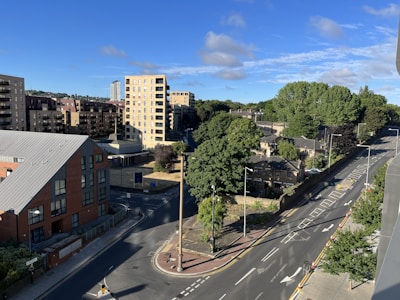 An urban landscape with residential buildings and a road. The area features several apartment complexes with multiple floors, surrounded by lush green trees. The sky is clear with a few clouds, indicating sunny weather. Streets are visible with marked lanes and some traffic cones, suggesting roadworks or restrictions. The scene is calm with no visible people or vehicles.