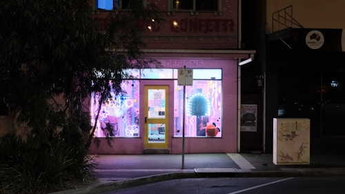 A storefront with a glowing pink and purple window display catches attention in an otherwise dark street. The design inside the window includes various shapes and neon lights, with reflections on the glass. A yellow door provides a contrasting focal point. A parking sign is visible in front of the store, and a tree partially obscures part of the shopfront.