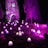 A whimsical scene illuminated at night features a collection of glowing purple mushrooms scattered on the forest floor. Among the towering tree trunks, two woven sculptures resembling organic creatures add an element of mystery and fantasy. A large, elaborate structure woven in a similar style stands prominently, casting a warm light from its windows.