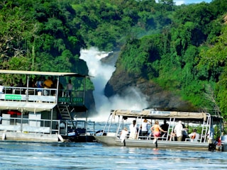 A scenic river scene with two boats filled with people observing a large waterfall surrounded by lush green vegetation. The waterfall cascades down rocky cliffs, creating mist and adding a sense of motion to the tranquil setting. The boats are labeled as 'African Queen' and are positioned on the calm water, reflecting the vibrant colors around them.
