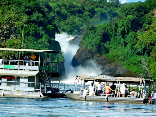 A scenic river scene with two boats filled with people observing a large waterfall surrounded by lush green vegetation. The waterfall cascades down rocky cliffs, creating mist and adding a sense of motion to the tranquil setting. The boats are labeled as 'African Queen' and are positioned on the calm water, reflecting the vibrant colors around them.