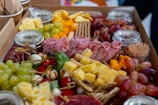 An assortment of charcuterie items artfully arranged in a box, featuring a variety of cheeses, cured meats, grapes, cherry tomatoes, leafy greens, and small skewers. Several jars are seen in the background, possibly containing dips or condiments.