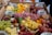 An assortment of charcuterie items artfully arranged in a box, featuring a variety of cheeses, cured meats, grapes, cherry tomatoes, leafy greens, and small skewers. Several jars are seen in the background, possibly containing dips or condiments.