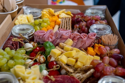 An assortment of charcuterie items artfully arranged in a box, featuring a variety of cheeses, cured meats, grapes, cherry tomatoes, leafy greens, and small skewers. Several jars are seen in the background, possibly containing dips or condiments.