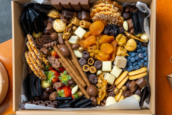 A box filled with a variety of snacks, including chocolates, dried fruits, nuts, fresh strawberries, wafers, and pretzels. The assortment is colorful and arranged neatly, featuring different textures and shapes.