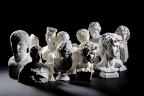 A collection of white classical busts arranged closely together on a dark background. The sculptures represent various figures with intricate hairstyles and expressions, showcasing a high level of detail in the craftsmanship.
