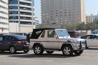 A silver SUV with a black soft top roof is parked in an urban parking lot. The vehicle is positioned in front of a dark sedan and is surrounded by several other parked cars. In the background, there are tall buildings with a modern architectural design, featuring many windows. The setting suggests a busy city environment with clear skies and light reflecting off the cars.