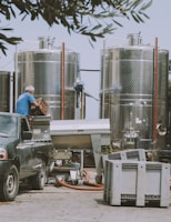 An outdoor industrial setting featuring two large metallic tanks. A person wearing a blue shirt is seen handling crates next to a dark-colored pickup truck. Various equipment and hoses are scattered around, and the scene is framed by overhanging tree branches.