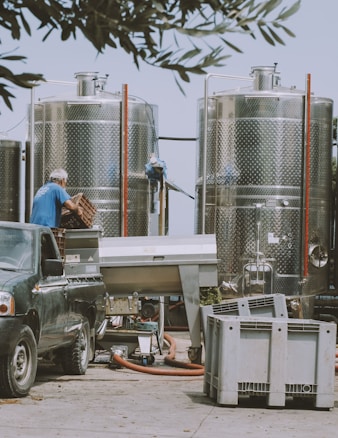 An outdoor industrial setting featuring two large metallic tanks. A person wearing a blue shirt is seen handling crates next to a dark-colored pickup truck. Various equipment and hoses are scattered around, and the scene is framed by overhanging tree branches.