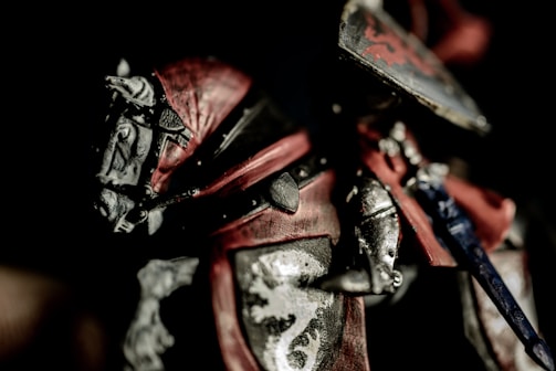 A detailed close-up of a medieval knight's armor, featuring intricate metalwork and vibrant red accents. The armor is ornately designed with heraldic symbols, including a lion motif on the breastplate.