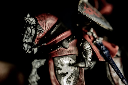 A detailed close-up of a medieval knight's armor, featuring intricate metalwork and vibrant red accents. The armor is ornately designed with heraldic symbols, including a lion motif on the breastplate.