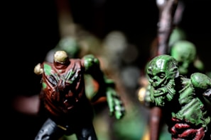 A close-up of two detailed, miniature zombie figures with grotesque features. The left figure has a headless appearance and is painted in dark colors with prominent textures, while the right figure has a skull-like face, green skin, and an exaggerated expression.