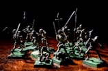 A group of miniature skeleton figures is positioned on a wooden surface. These figures are intricately detailed, each holding various types of weapons such as swords and spears. They stand on green square bases, giving them a battle-ready appearance. The lighting creates dramatic shadows, emphasizing the eerie and menacing nature of the scene.