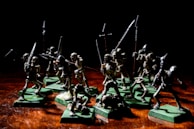 A group of miniature skeleton figures is positioned on a wooden surface. These figures are intricately detailed, each holding various types of weapons such as swords and spears. They stand on green square bases, giving them a battle-ready appearance. The lighting creates dramatic shadows, emphasizing the eerie and menacing nature of the scene.