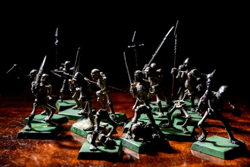 A group of miniature skeleton figures is positioned on a wooden surface. These figures are intricately detailed, each holding various types of weapons such as swords and spears. They stand on green square bases, giving them a battle-ready appearance. The lighting creates dramatic shadows, emphasizing the eerie and menacing nature of the scene.