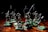 A group of miniature skeleton figures is positioned on a wooden surface. These figures are intricately detailed, each holding various types of weapons such as swords and spears. They stand on green square bases, giving them a battle-ready appearance. The lighting creates dramatic shadows, emphasizing the eerie and menacing nature of the scene.